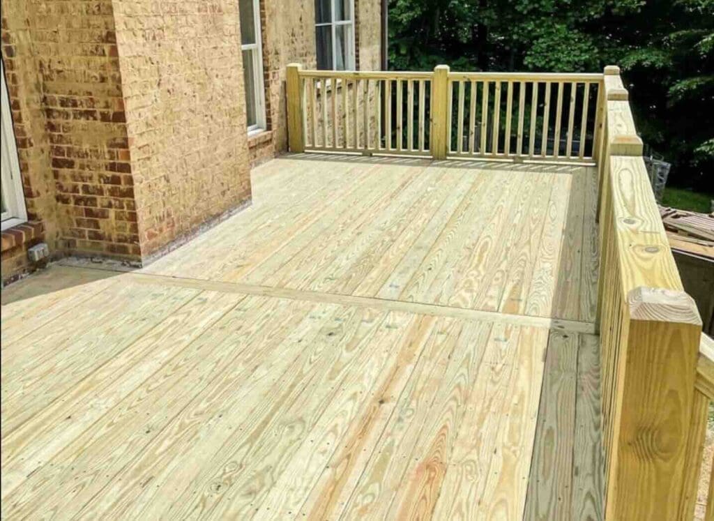 Newly constructed wooden deck with railing, showcasing clean, unfinished boards, adjacent to brick wall of a house, emphasizing outdoor living space enhancement and deck maintenance.